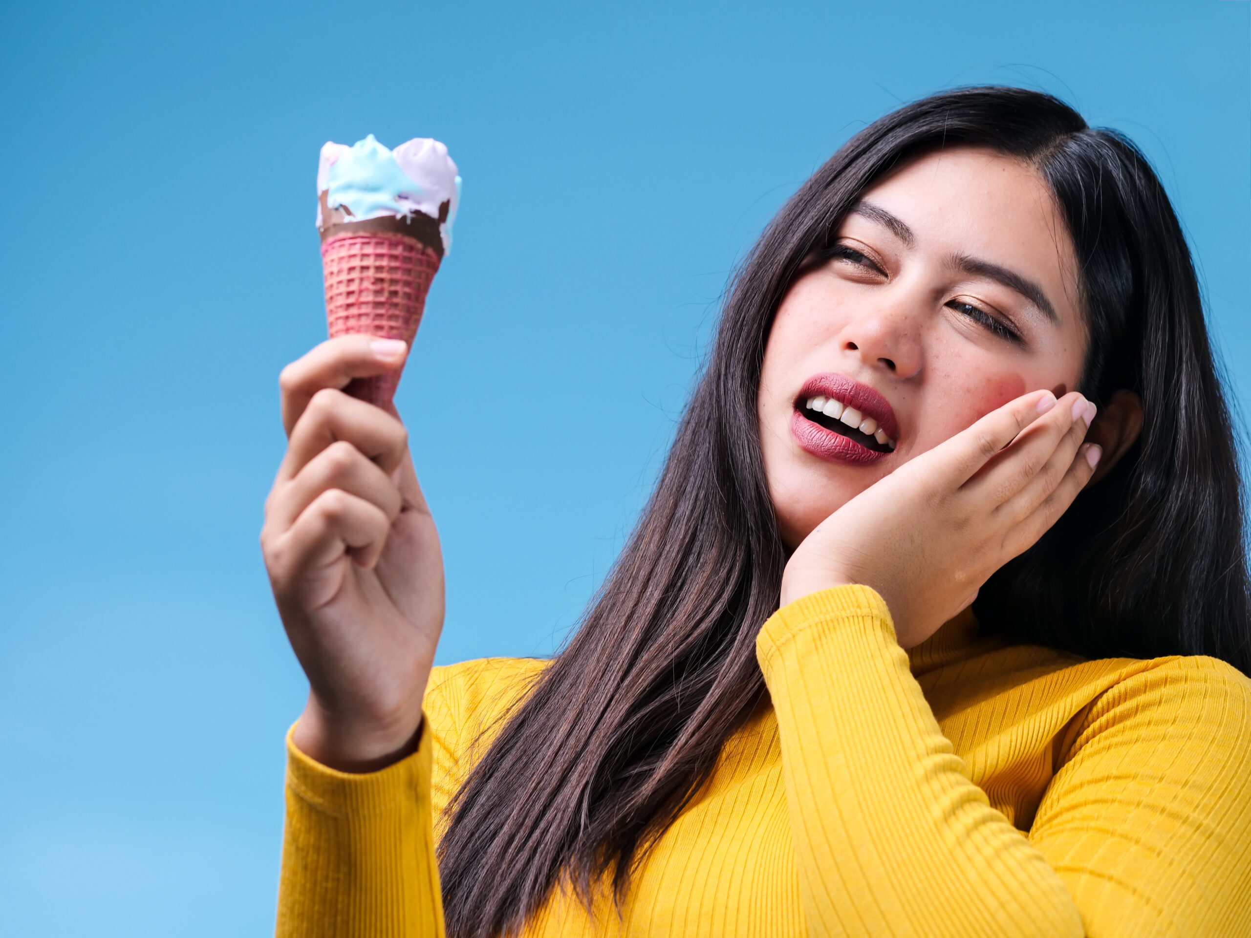 Young Asian woman with sensitive teeth and cold ice cream on color background